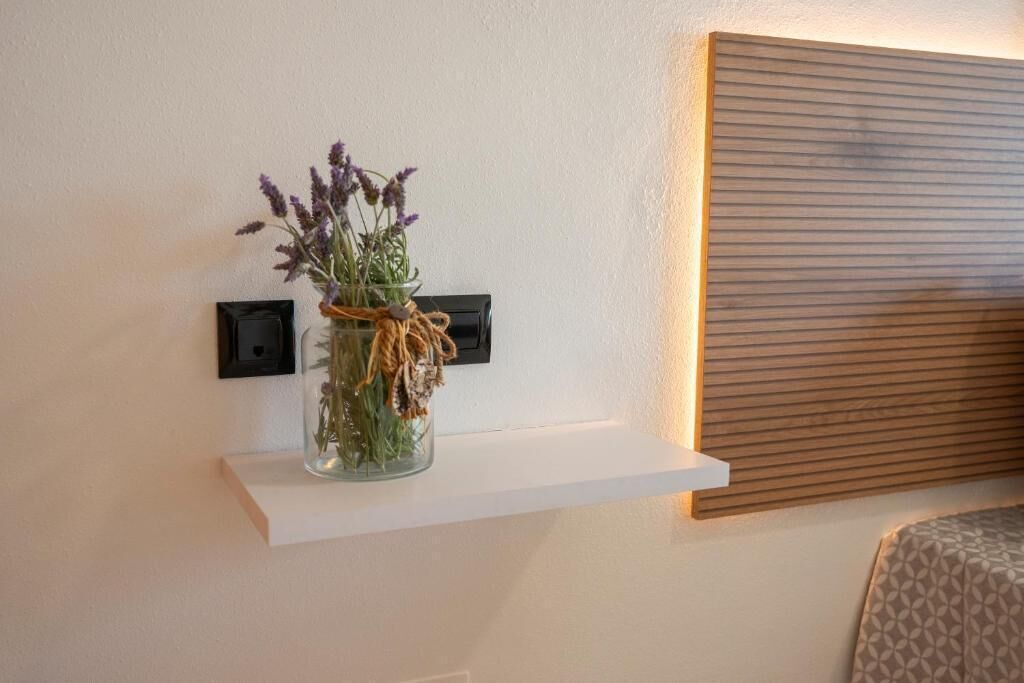 A white shelf with a glass vase containing dried flowers and greenery, mounted on a wall with a power outlet and a wooden panel.