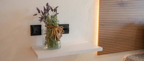 A white shelf with a glass vase containing dried flowers and greenery, mounted on a wall with a power outlet and a wooden panel.