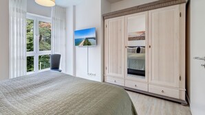 Une chambre avec un grand lit, une armoire, un téléviseur fixé au mur et une fenêtre avec des rideaux.