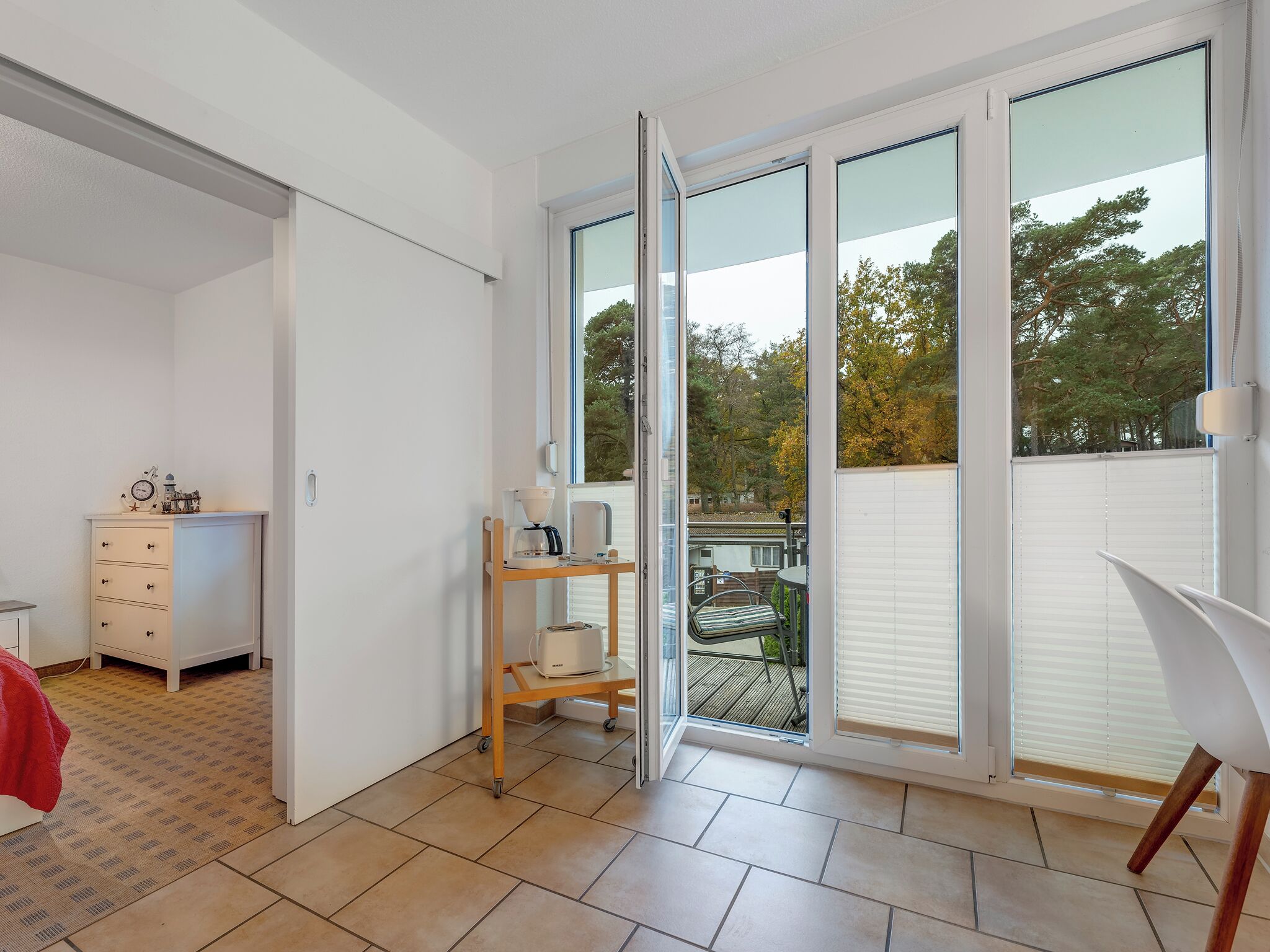 A modern room with a sliding glass door leading to a balcony, a white dresser, a coffee machine, and a chair.