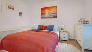 A bedroom with a large bed, a nightstand, a dresser, and a painting of a sunset over the ocean. - Haus Eichhorst, Whg. 03: Close to the sandy beach in Trassenheide (Trassenheide)