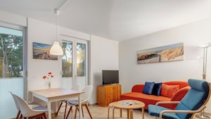 A modern living room with a wooden dining table, white chairs, a red sofa with blue cushions, a wooden cabinet, and a TV. - Haus Eichhorst, Whg. 03: Close to the sandy beach in Trassenheide (Trassenheide)