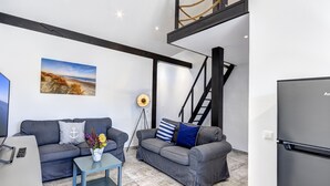 A modern living room with two grey sofas, a coffee table, a flat-screen TV, a refrigerator, and a painting of a beach scene. - Emilia vacation apartment in Trassenheide: WiFi, garden & only 1.9 km to the beach (Trassenheide)
