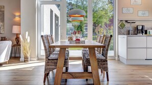 Un coin repas avec une table en bois et des chaises en osier, de grandes fenêtres et une vue sur la verdure environnante.