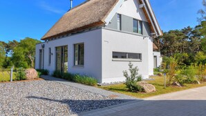 Une maison moderne aux murs blancs, dotée d’un toit de chaume, de grandes fenêtres et d’une allée gravillonnée.
