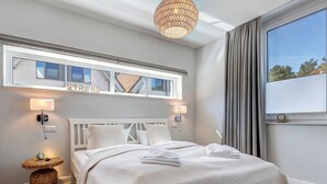 Une chambre moderne avec un grand lit, deux lampes de chevet, une table d’appoint en osier et une vue sur l’extérieur grâce à une grande fenêtre.