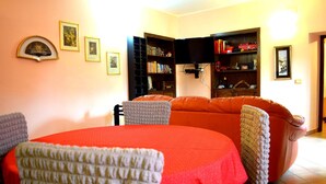 Ein Essbereich mit einem roten Tisch, grauen Stühlen und einer Couch. Im Hintergrund befinden sich Regale und ein Fernseher.