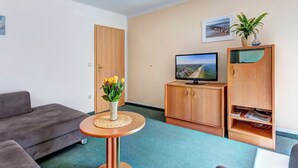 A modern living room with a grey sofa, a small wooden table with a vase of flowers, a television on a wooden cabinet, and a bookshelf. - Hansehus, Whg. 5: close to the beach in Zinnowitz with balcony and WLAN (Zinnowitz)