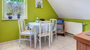 Een eetgedeelte met een tafel voor twee personen, stoelen, een raam met gordijnen, een radiator en een klein plankje met een broodrooster.