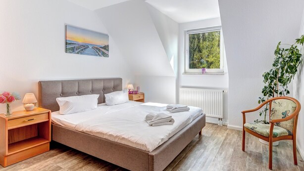 Een slaapkamer met een groot bed, een nachtkastje, een stoel, een raam met uitzicht op bomen en een schilderij aan de muur.
