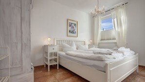 A white bedroom with a bed, nightstands, a dresser, and a window with curtains.