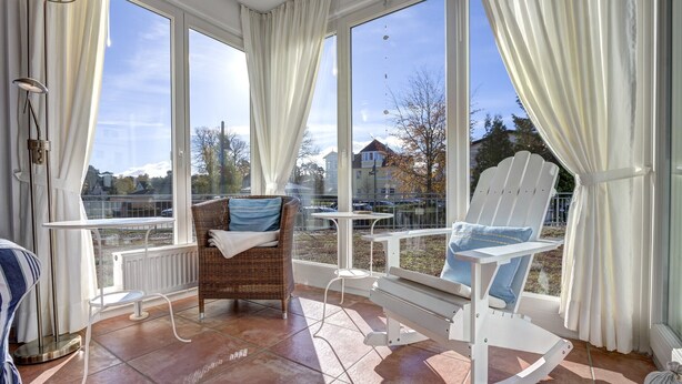 A sunlit room with a large window, a wicker chair, a rocking chair, and a small table.
