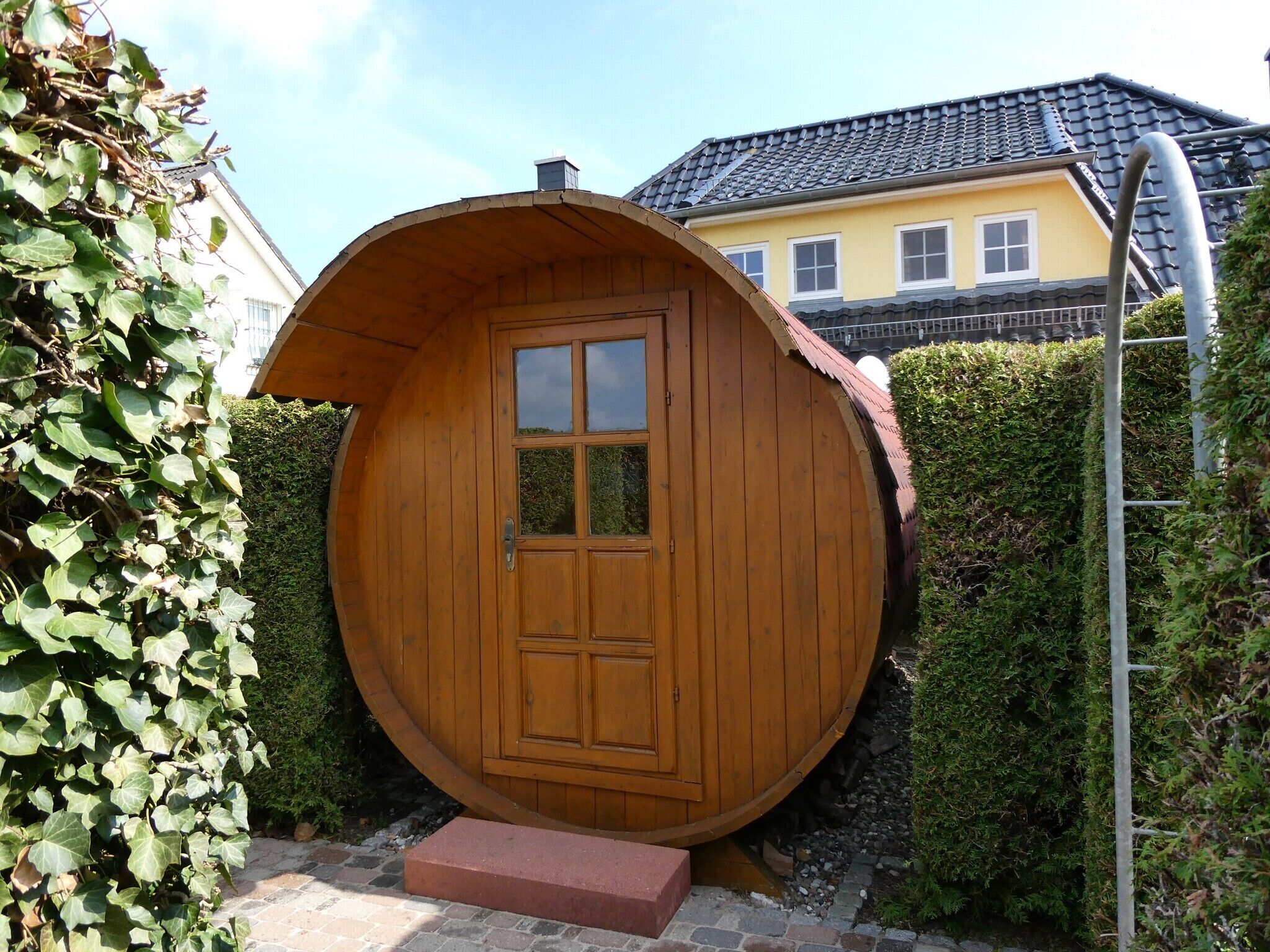 Una estructura circular de madera con puerta y ventanas, rodeada de follaje y un área pavimentada.