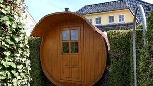 Uma estrutura circular de madeira com porta e janelas, rodeada por vegetação e um espaço pavimentado.