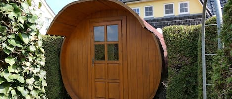 A wooden, circular structure with a door and windows, surrounded by greenery and a paved area.
