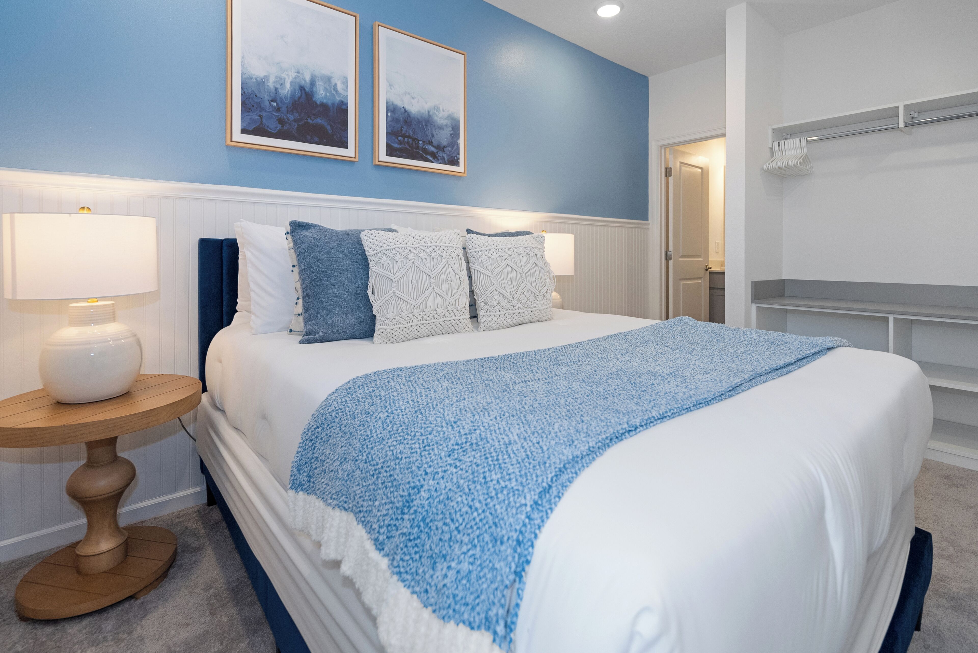 Ein Schlafzimmer mit einem Bett, zwei Nachttischlampen, einem runden Holztisch und einer blauen Wand mit zwei gerahmten Bildern.