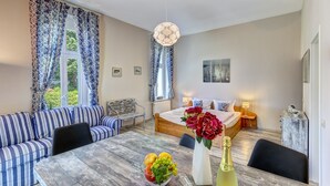 A modern bedroom with a wooden bed, a sofa, a dining table with fruit and a bottle, and a view of greenery outside. - Vacation apartment "Küstenwald" in the Zinnowitzer Hof on Usedom! (Zinnowitz)