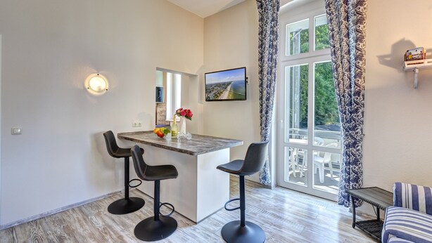 A modern kitchen with a central island, two bar stools, a dining table, and a large window with patterned curtains. - Vacation apartment "Küstenwald" in the Zinnowitzer Hof on Usedom! (Zinnowitz)