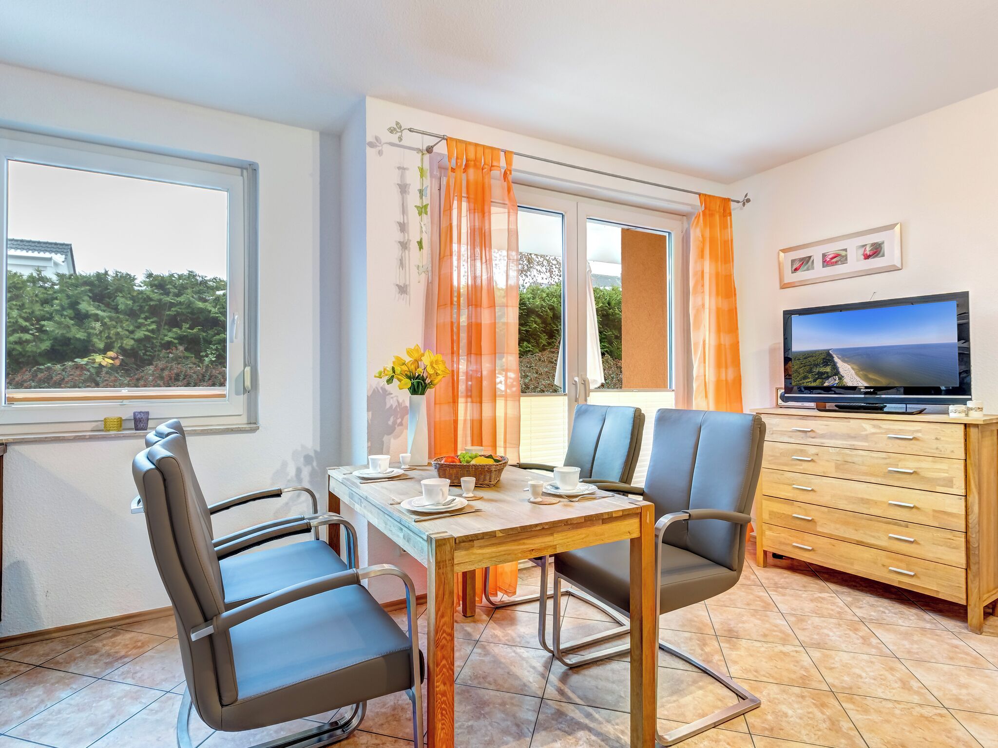 A dining area with a table set for two, a television on a wooden cabinet, and a window with curtains.