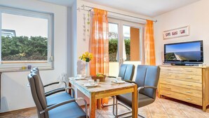 Un comedor con una mesa dispuesta para dos personas, un televisor sobre un mueble de madera y una ventana con cortinas.