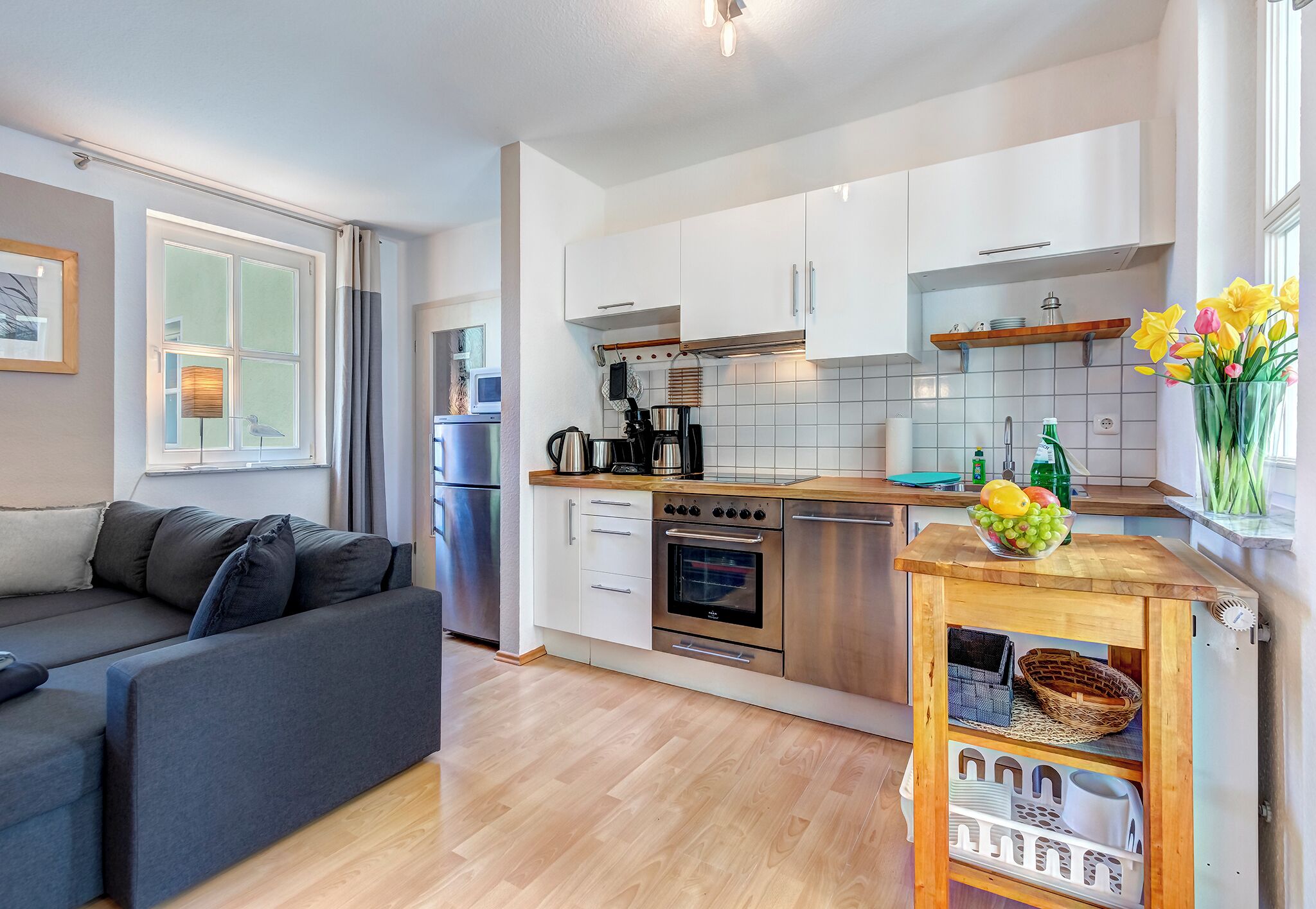 Una cocina moderna con armarios blancos, una encimera de madera y un horno empotrado. Hay una pequeña zona de comedor con una mesa y sillas, y un sofá en la sala de estar contigua.