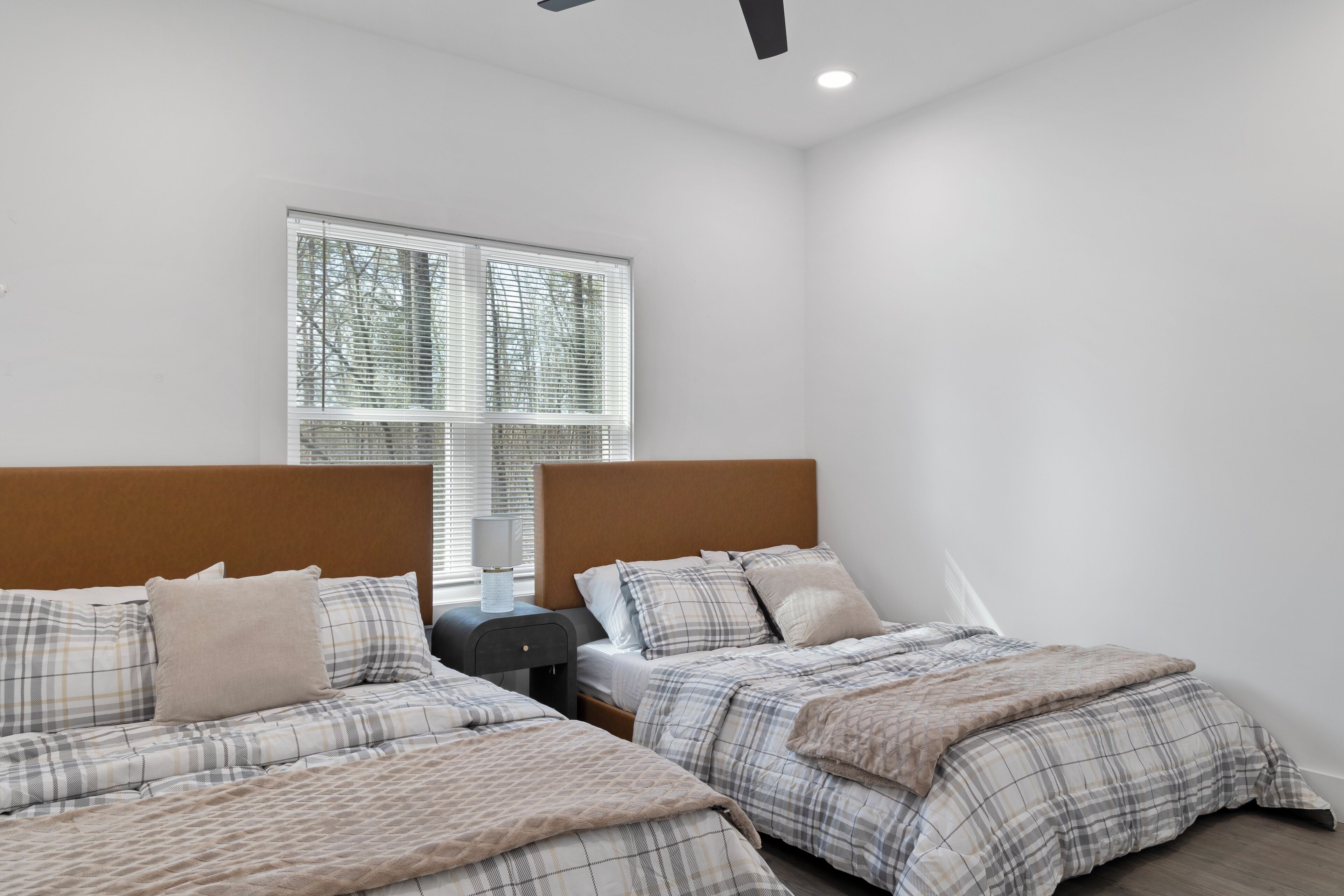 A bedroom with two beds, each with a headboard, a nightstand, and a lamp. There is a window with blinds and a view of trees outside.