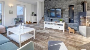 Una sala de estar moderna con pared de piedra, un televisor de pantalla plana, un mueble blanco para equipo audiovisual y una zona de comedor con mesa y bancos blancos.