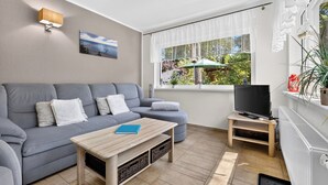 Ein modernes Wohnzimmer mit einem grauen Sofa, einem Couchtisch, einem Fernseher auf einem Lowboard und einem Fenster mit Blick ins Grüne.
