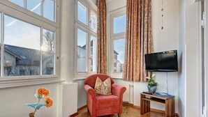 Ein Wohnzimmer mit einem roten Sessel, einem kleinen Tisch mit Blumenvase und einem an der Wand montierten Fernseher.