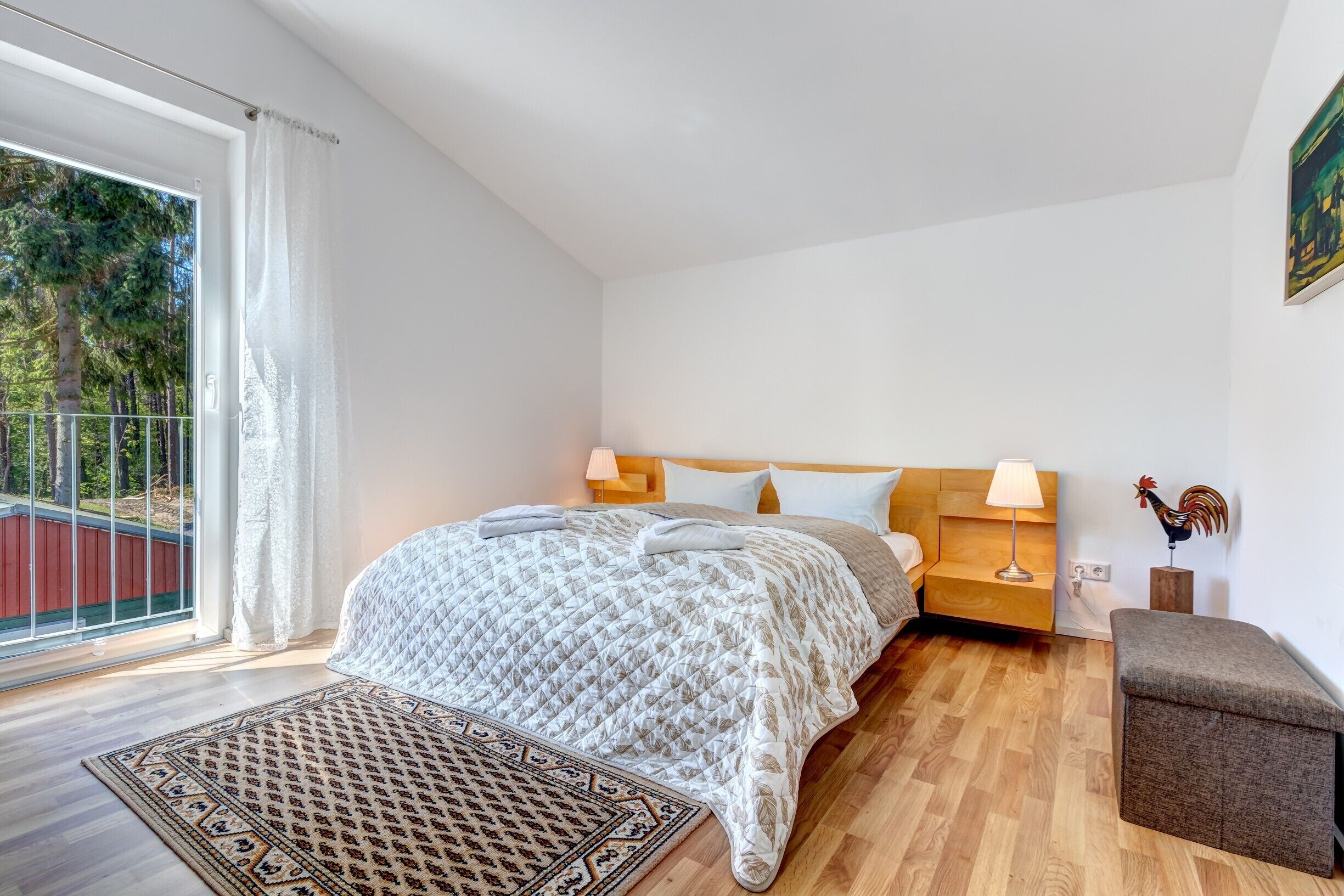 Quarto com uma cama grande, uma mesa de cabeceira com abajur, um banco de madeira e vista para árvores por uma porta de correr.