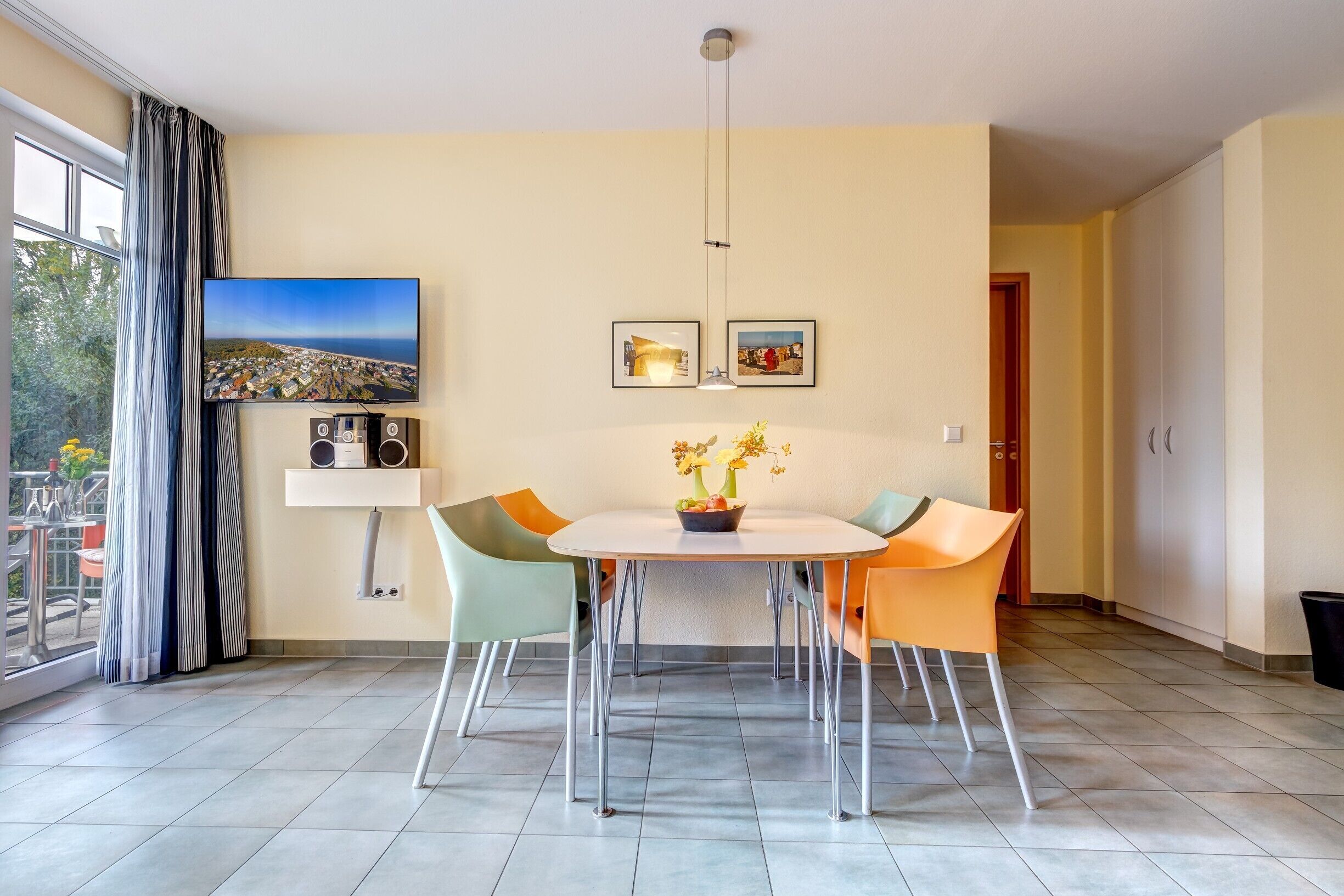 Un coin repas avec une table ronde, quatre chaises, un vase avec des fleurs, un téléviseur à écran plat, deux tableaux encadrés et un haut-parleur posé sur une étagère.