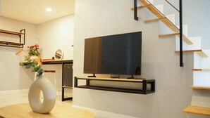 A modern living room with a flat-screen TV mounted on a black shelf, a round wooden table with a white vase, and a staircase with wooden steps and black railings. - Naoat Resort Farm & Cafe (Mae Taeng)