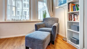 Uma sala de estar moderna com uma poltrona cinza e apoio para os pés, uma estante com livros e objetos decorativos, e uma janela grande com cortinas leves.