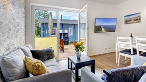 A modern living room with a grey sofa, a small table with a vase of flowers, a white dining table with chairs, and a TV mounted on the wall.