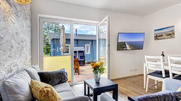 Ein modernes Wohnzimmer mit grauem Sofa, einem kleinen Tisch mit Blumenvase, einem weißen Esstisch mit Stühlen und einem an der Wand montierten Fernseher.