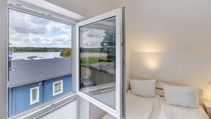 A hotel room with a large window, a bed with white bedding, and a view of a blue building and a body of water.