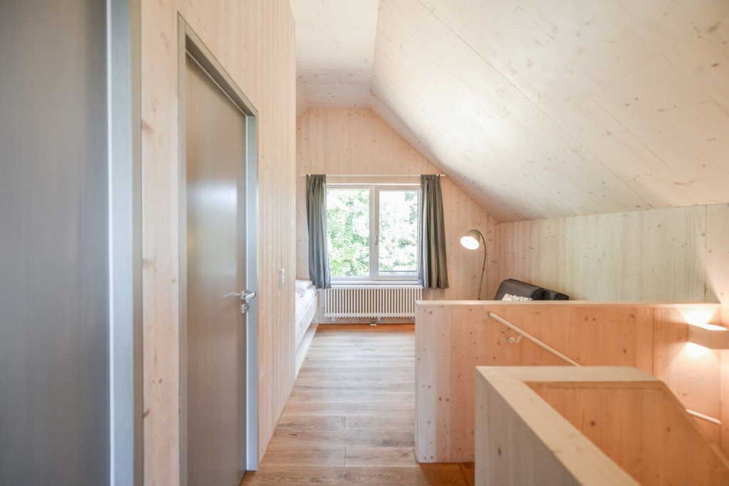 En smal trægang fører hen til et vindue med udsigt over grønne områder, en radiator og et lille skrivebord med lampe.