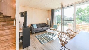 A modern interior with a wooden floor, a grey sofa with blue cushions, a black heater, wicker chairs, and a large window offering a view of greenery. - "Söt Mine" Seaside Cottage – a stylish retreat on the Baltic Sea for up to 7 people (Loddin OT Stubbenfelde)