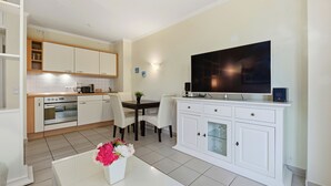 Een moderne keuken met witte kasten, een eethoek met een tafel en stoelen, een flatscreen-tv op een wit kastje en een vaas met bloemen op een witte tafel.