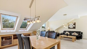 Una sala de estar moderna con comedor, que incluye una mesa y sillas de madera, un sofá negro y un amplio ventanal con vistas a árboles.