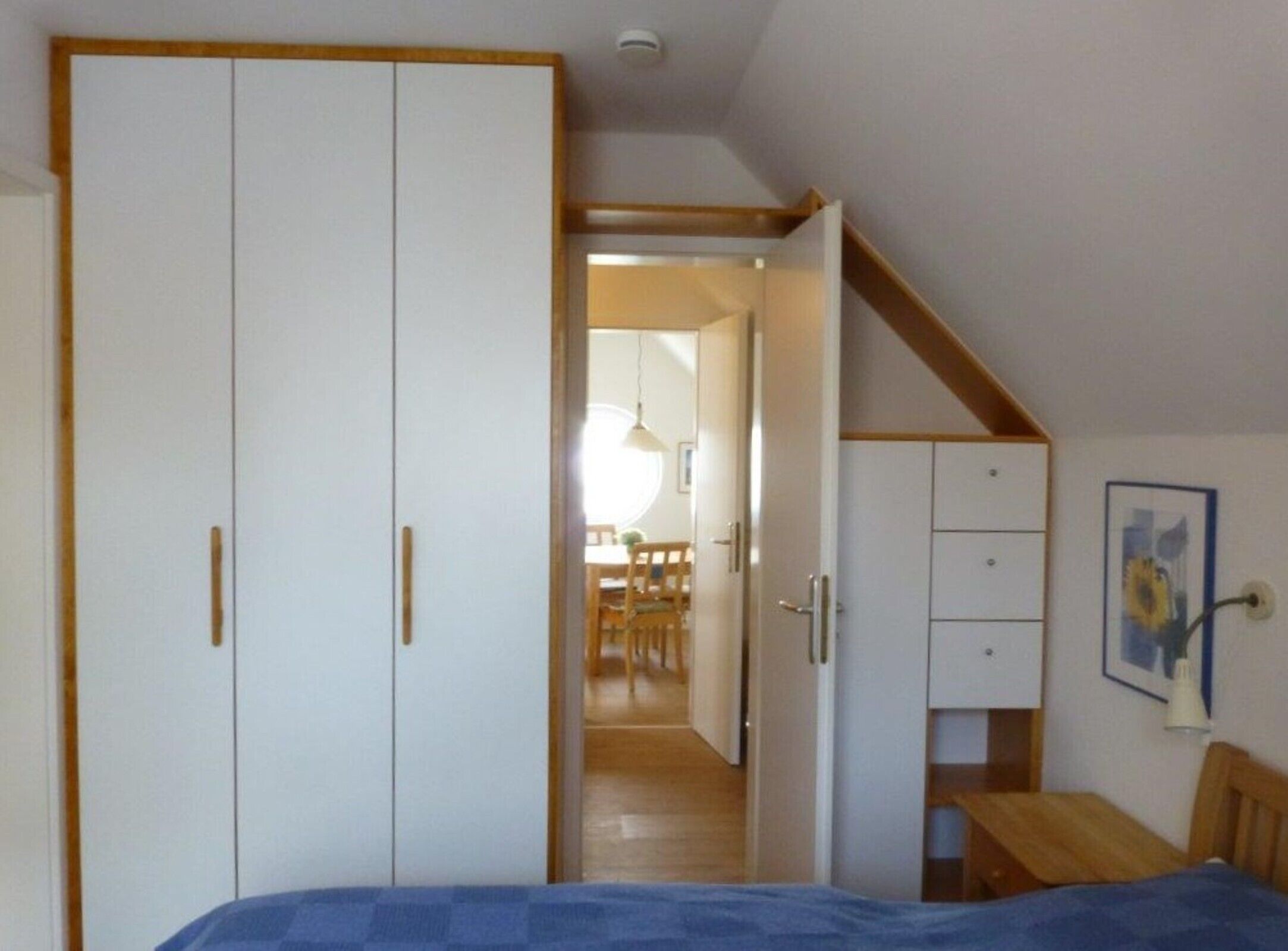 A bedroom with a bed, a wardrobe, a desk, and a chair. There is a hallway leading to a dining area.