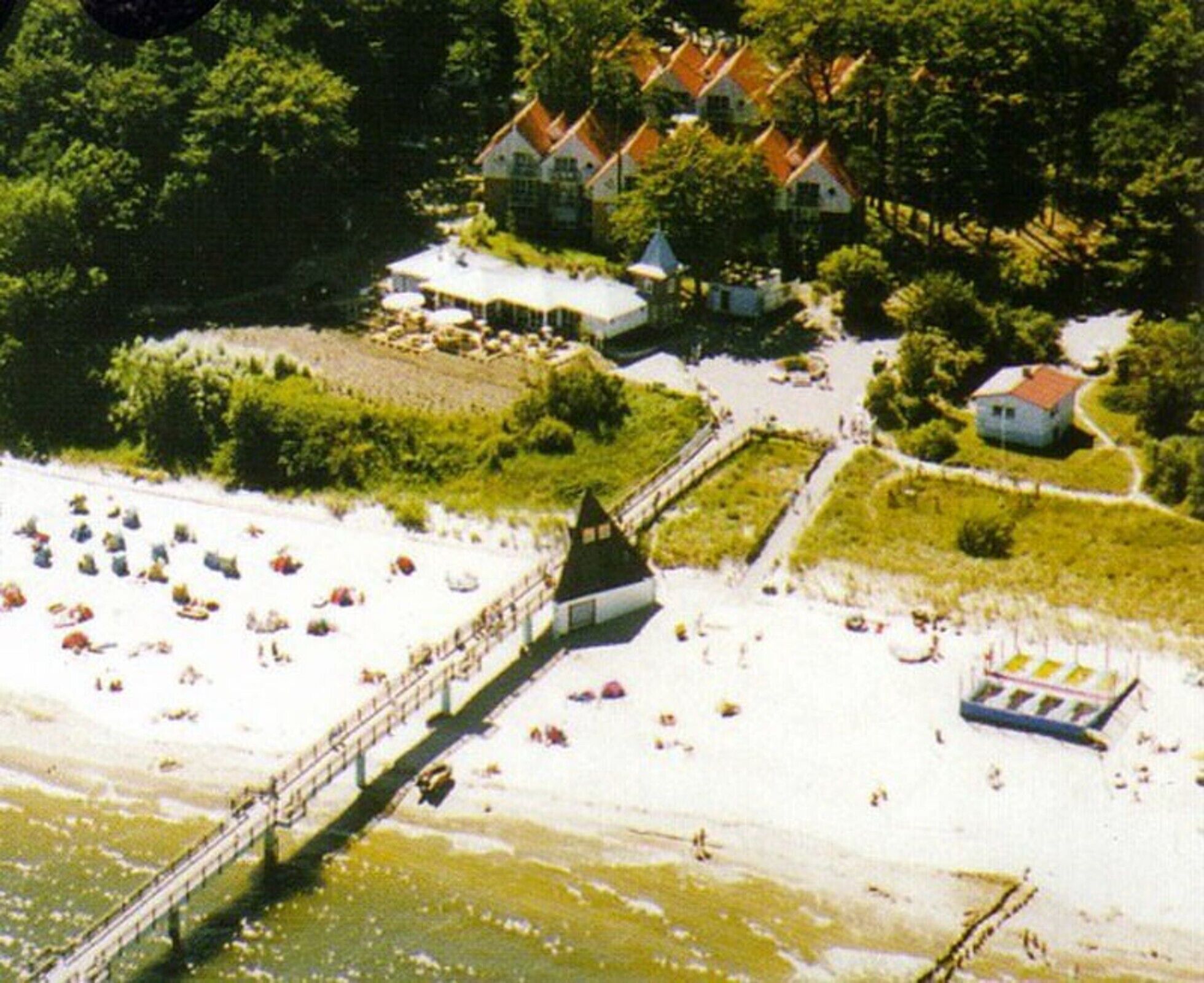 Vista aérea de uma praia com um píer, guarda-sóis e uma fileira de casas com telhados vermelhos.
