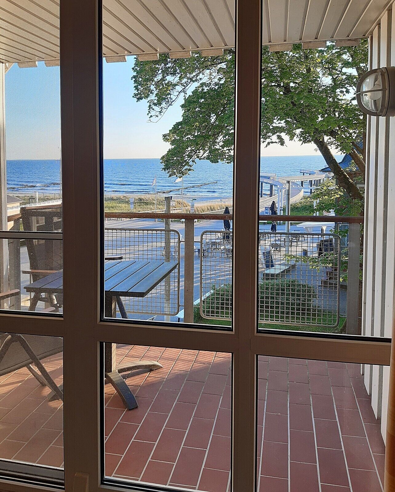 A view from a covered patio with a brick floor, overlooking a beach area with tables and chairs, and a clear view of the ocean.