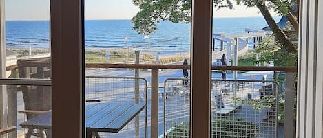 A view from a covered patio with a brick floor, overlooking a beach area with tables and chairs, and a clear view of the ocean.