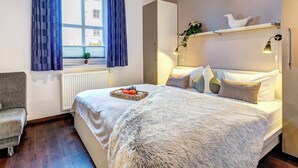 A bedroom with a large bed, a window with curtains, a radiator, a small plant, and a bedside table with a lamp.