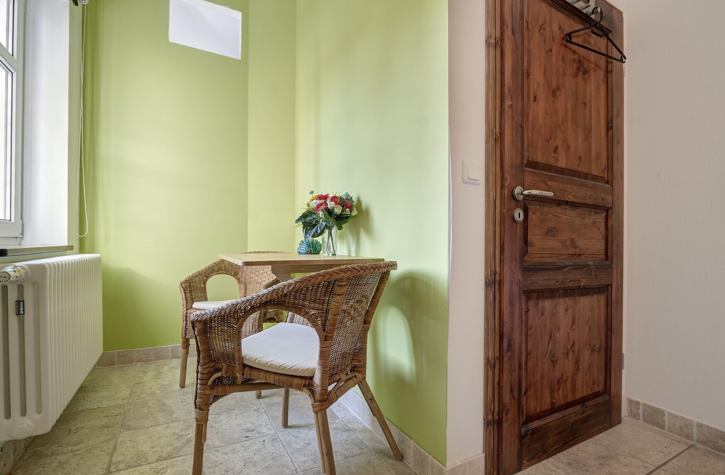 Una habitación con una pared verde, una puerta de madera, una silla de mimbre, una mesita con un jarrón de flores y un radiador.