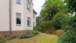 A building with a brick base and white upper walls, surrounded by a grassy yard and dense foliage.