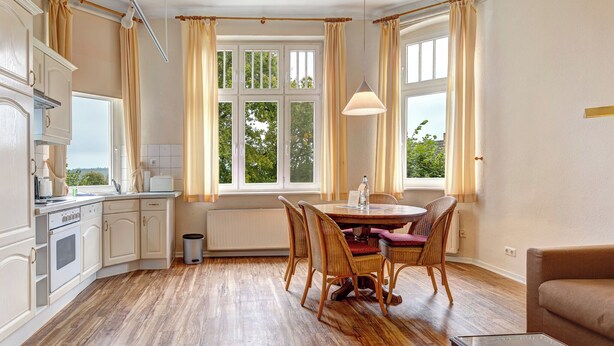 A kitchen with a dining area, wooden flooring, and large windows with curtains.