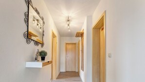 A hallway with a wooden floor, a white wall, a decorative mirror, and a small shelf with a plant.