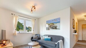 A modern living room with a grey sofa, a small round table, and a wooden side table with a lamp. There is a large window with curtains, a painting of birds on the wall, and a mirror above the door.